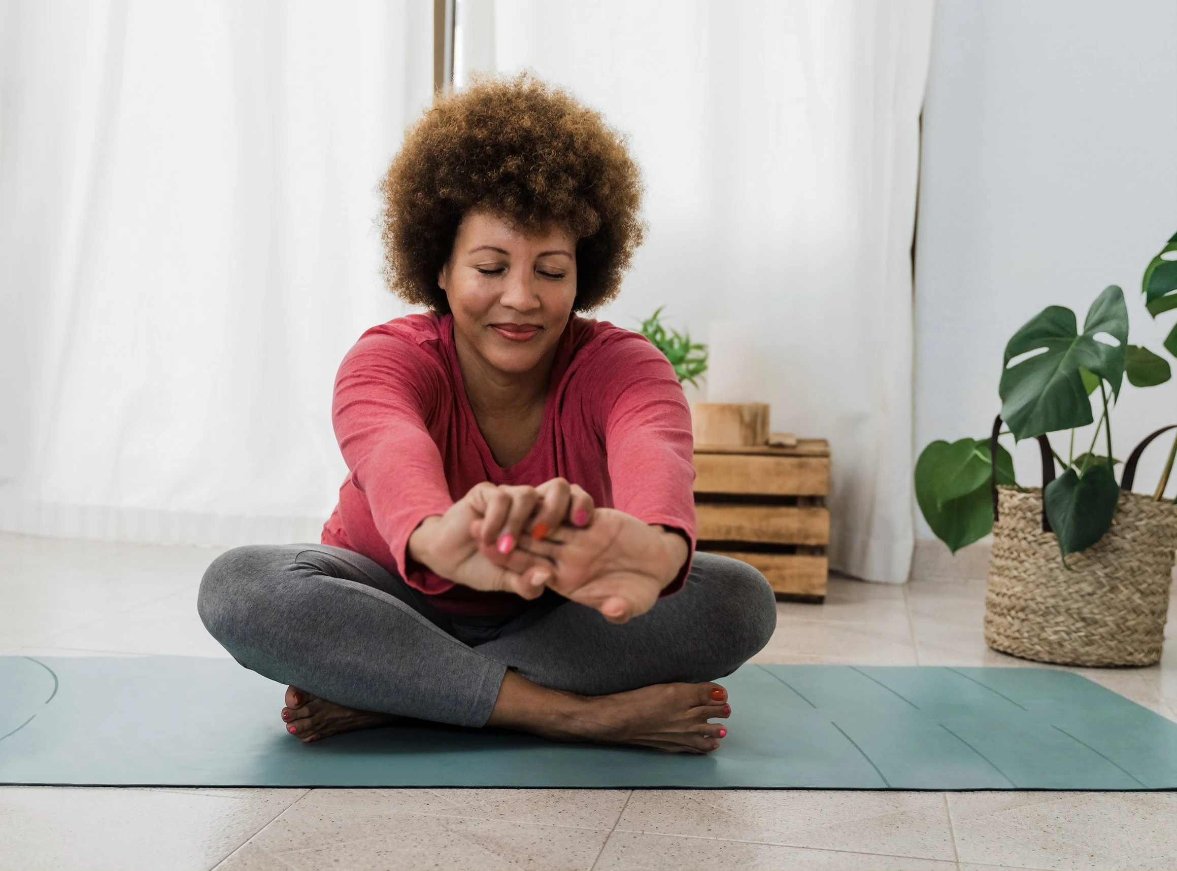A woman sits cross-legged on a yoga mat at home, stretching her arms forward